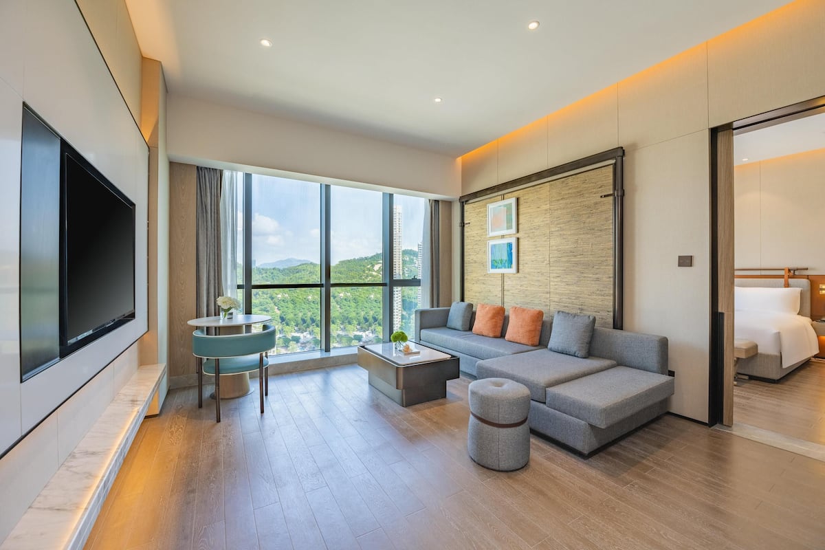 Ein modernes Wohnzimmer mit einem großen Flachbildfernseher, einem grauen Sofa mit orangefarbenen Kissen, einem runden Couchtisch und Blick auf Grünpflanzen durch große Fenster.