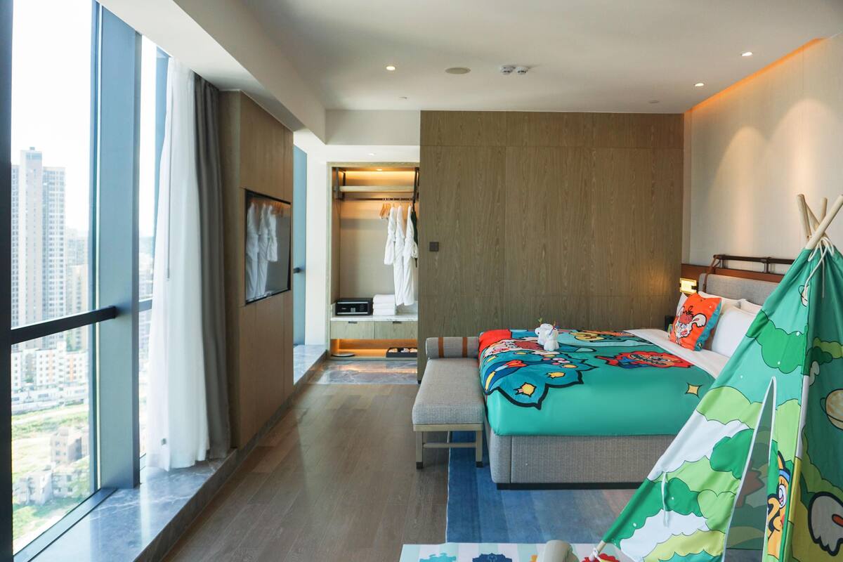 Une chambre d’hôtel moderne avec un grand lit, un banc, une vue sur la ville, une armoire et une décoration sur le thème des tipis.