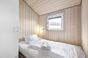A single bed with white linens and towels, a window with a view of a building, and wooden walls.
