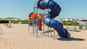 A playground with a blue spiral slide, a red and yellow climbing structure, and a picnic table on a sandy beach. - First Camp Lakolk strand- Römö (Rømø)