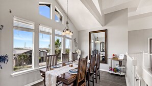 Un comedor con una mesa de madera, sillas y una araña. Hay ventanas grandes con persianas, un espejo y vistas al exterior.