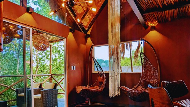 A spacious room with a wooden floor, large windows, and a thatched ceiling. There are wicker chairs and a hanging chair. - TreeHouse EcoChic/3 min walk beach/Wifi/AC/kitchen/King Size Bed (Isla Holbox)