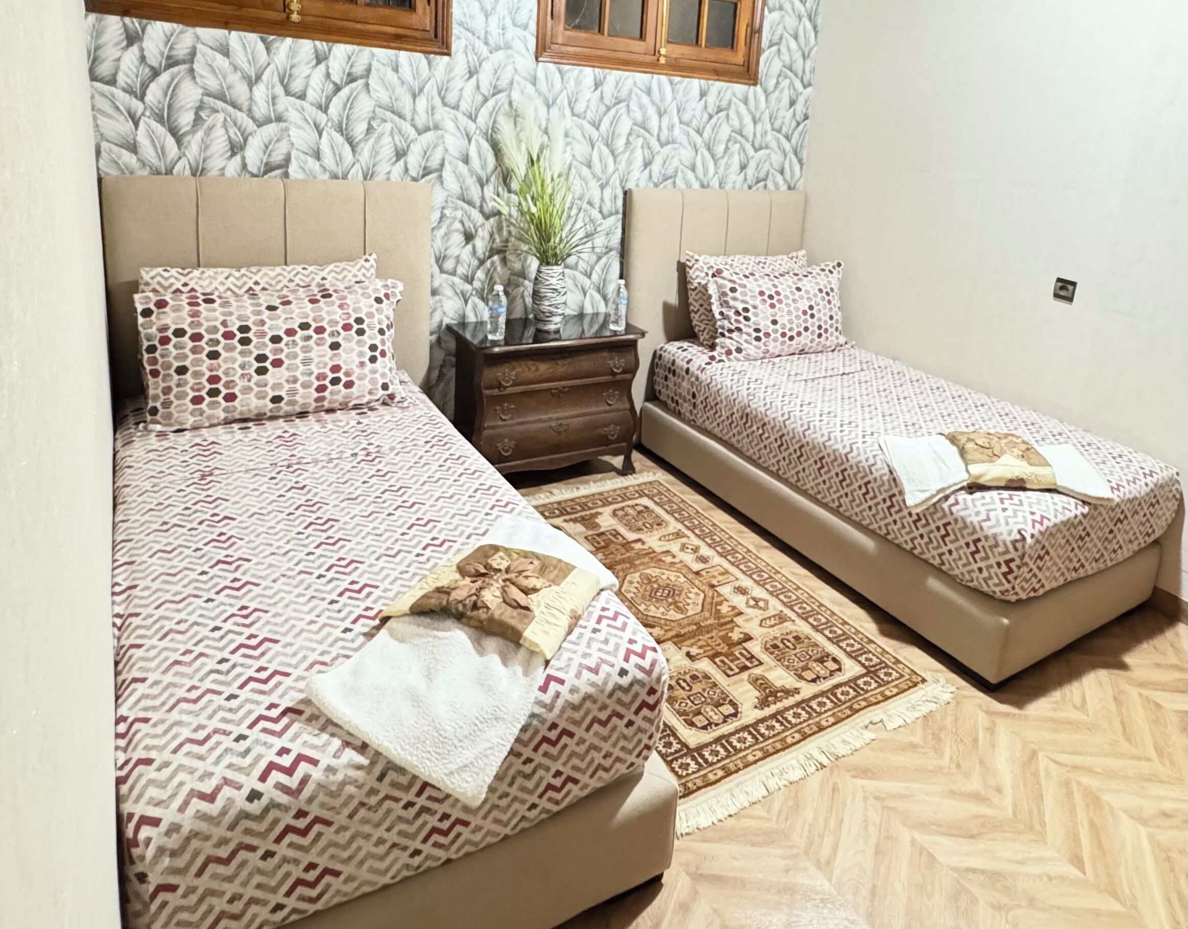 A bedroom with two beds, a wooden dresser, a patterned rug, and a wall with a leaf design.