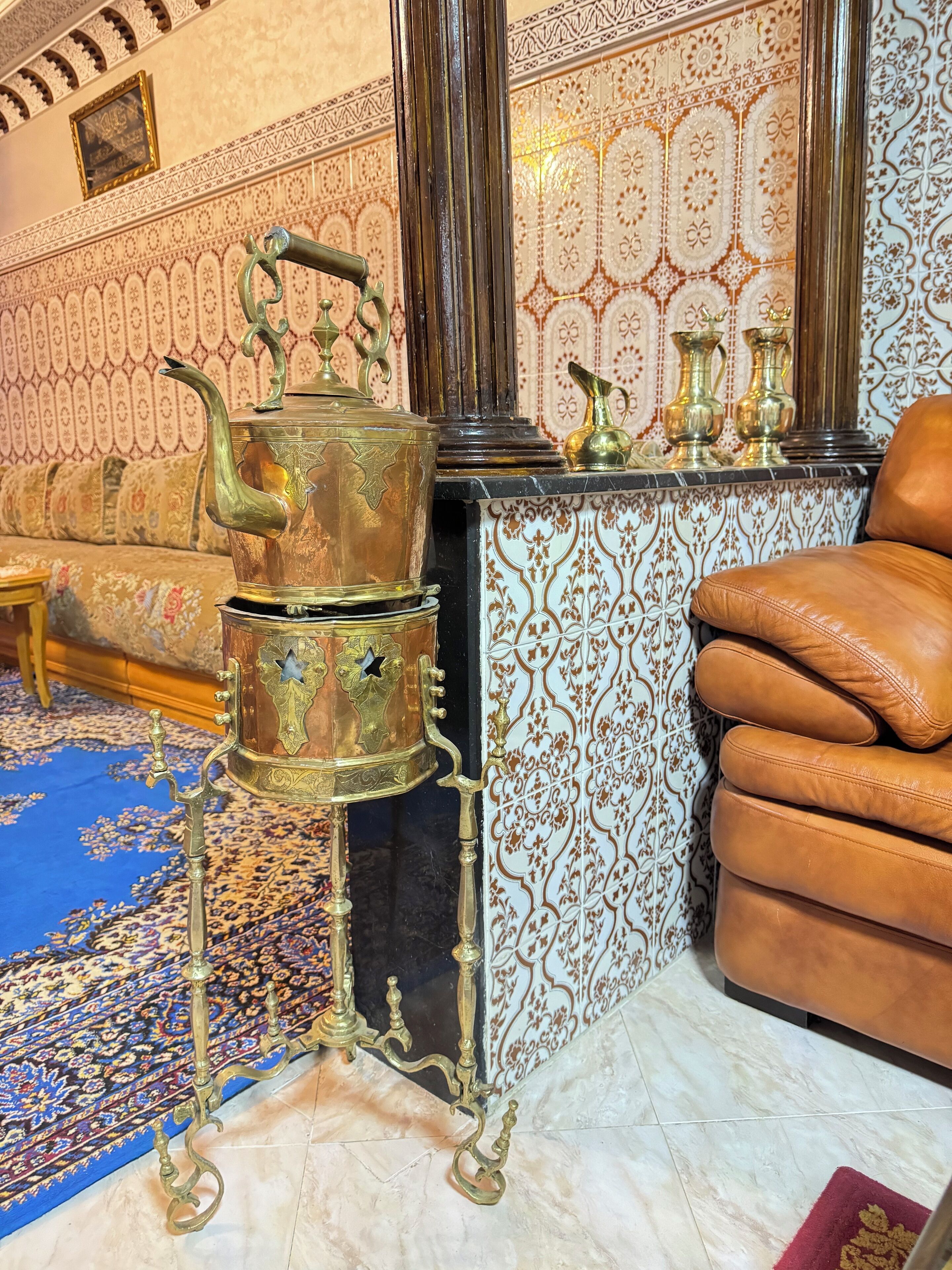 A traditional room with ornate wall tiles, a brass teapot on a stand, a sofa, and a leather armchair.