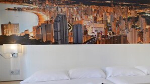 Hotellihuone, jossa on sänky, tyynyjä, yöpöytä lampun kanssa ja seinällä kaupunkimaisemaa esittävä seinämaalaus.