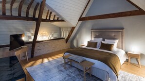 A bedroom with a sloped ceiling, a bed with multiple pillows, a bench, a TV, and a lamp. - VAU D'ARZ MANOR - Gîte LA GACILLY (MALANSAC)