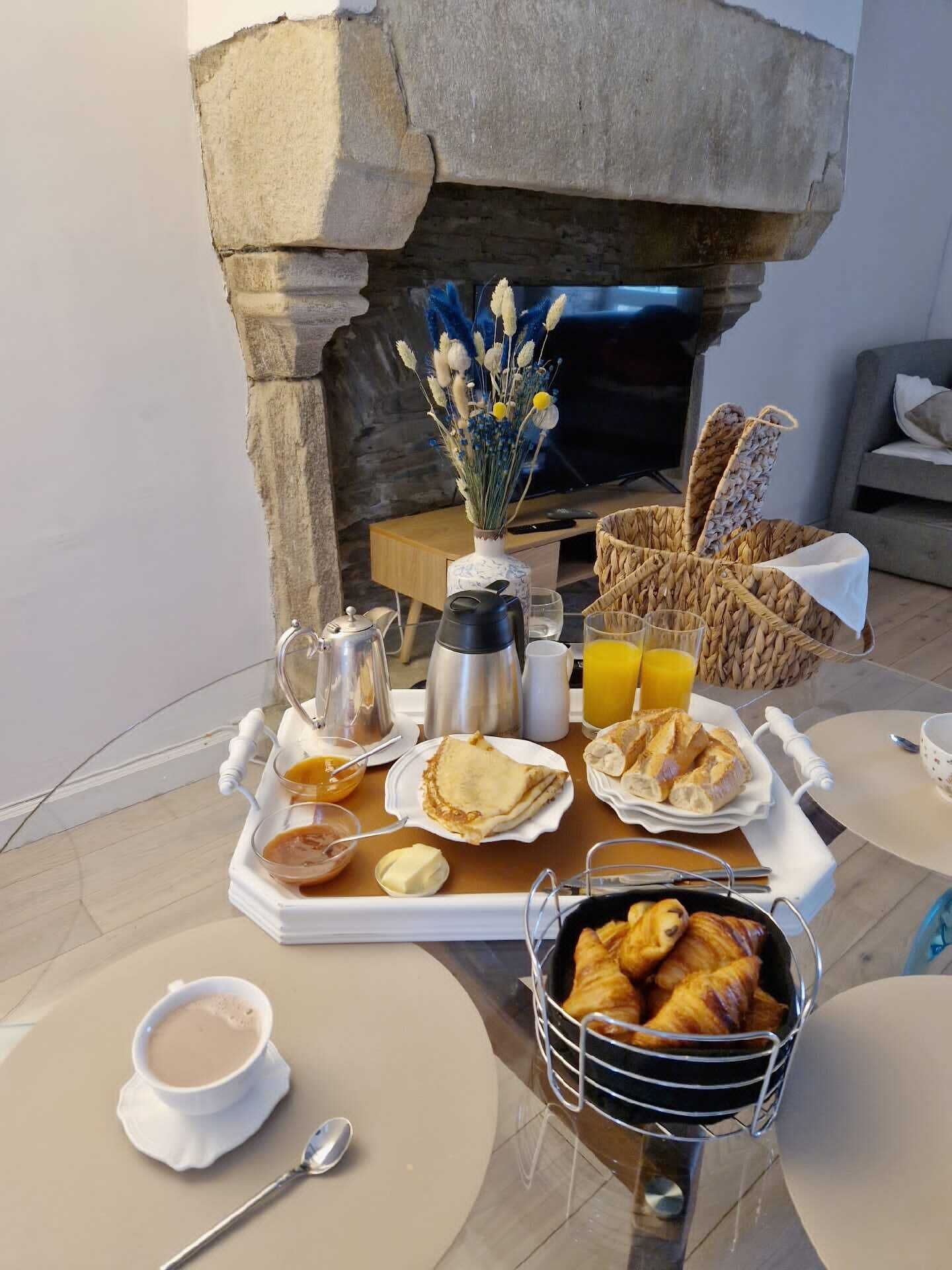 Un coin petit-déjeuner agrémenté d’une cheminée en pierre, d’un plateau avec des croissants, des pâtisseries, du café et du jus de fruits.