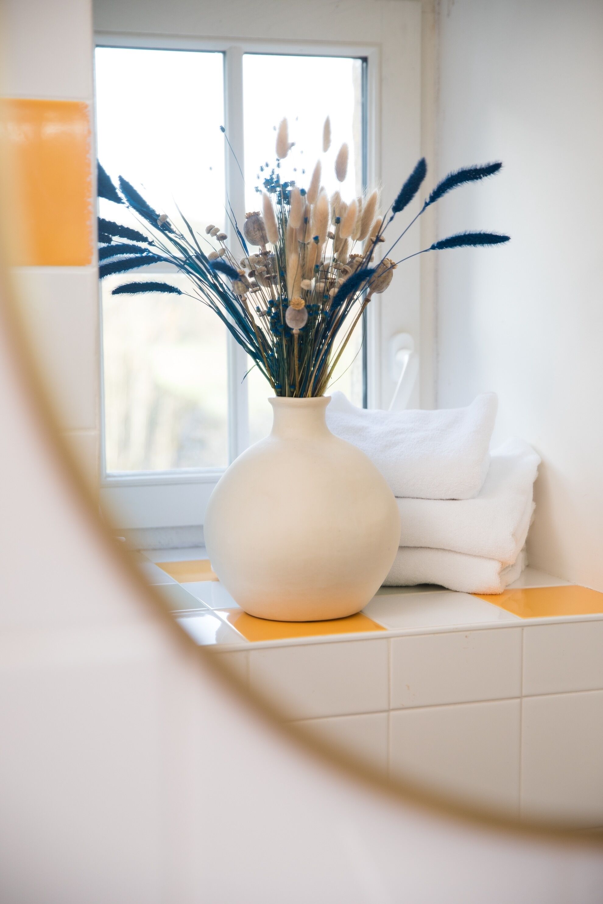 A white vase with dried plants on a white tiled surface near a window.