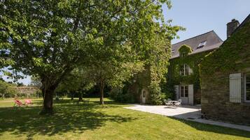A stone building with ivy-covered walls, a grassy courtyard with a table and chairs, and a large tree providing shade.