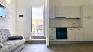 A modern kitchen with white cabinets and a built-in oven. There is a grey sofa and a small table with chairs in the living area.