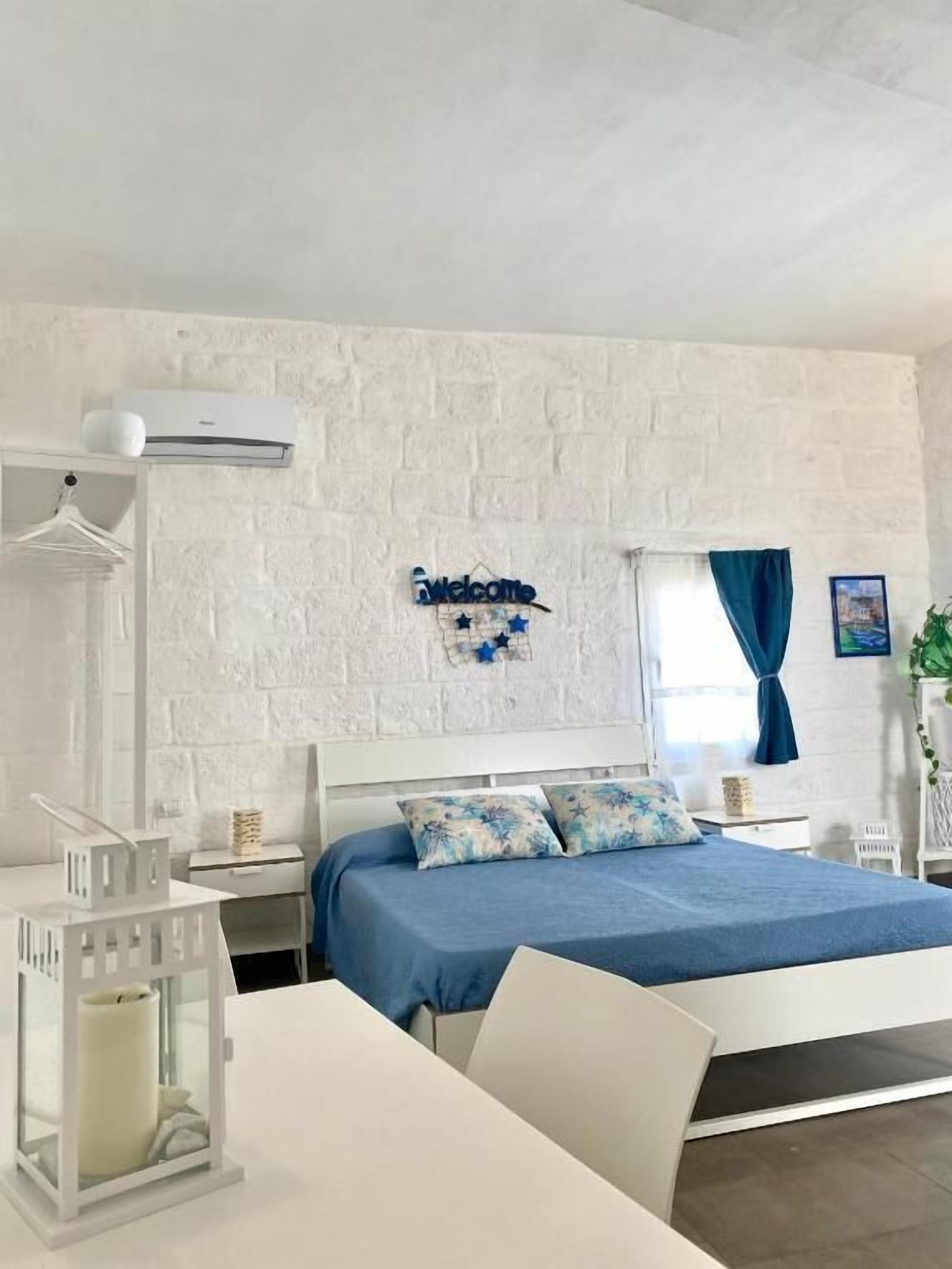 A bedroom with a white brick wall, a bed with blue bedding, a white bedside table, a hanging light fixture, and a 'Welcome' sign with stars.