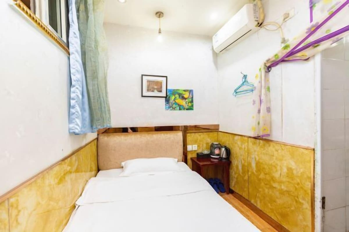 A small, compact hotel room with a bed, a small table with a kettle, and a wall-mounted air conditioner.
