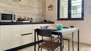 A modern kitchen with a microwave, kettle, and cabinets. There is a dining table with chairs and a window with blinds. - Allegria Pool House (Monopoli)