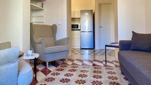 A compact living space with a sofa, armchair, and a small table. A kitchen area with a refrigerator and microwave is visible in the background. - Rouge (Monopoli)