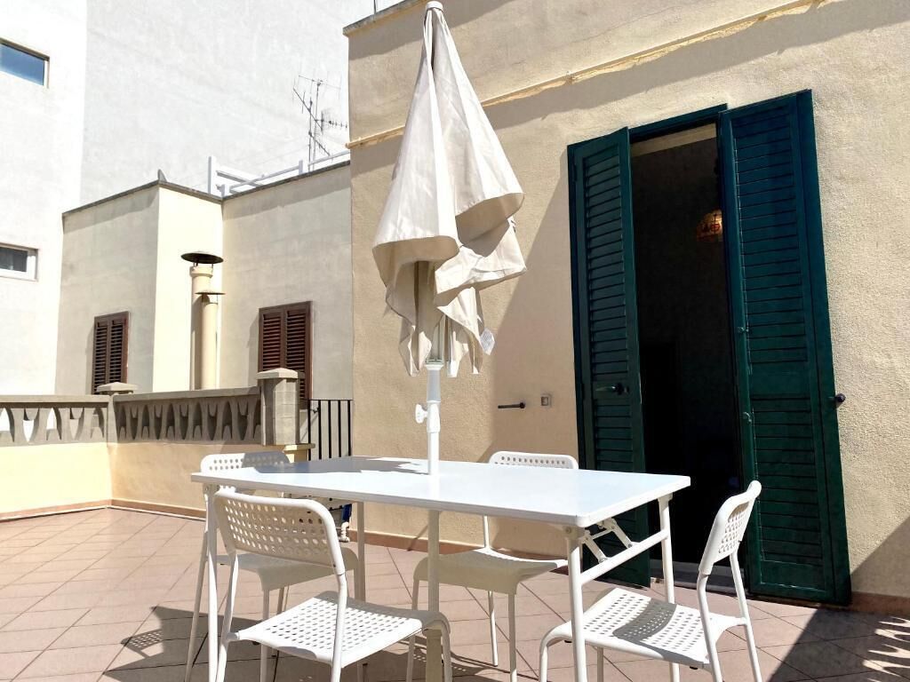 Une table et des chaises blanches sont disposées sur une terrasse carrelée, sous un parasol blanc.