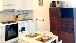Een kleine keuken met witte kastjes, een fornuis, oven, magnetron en spoelbak. Er is een eettafel met stoelen en een klok aan de muur.