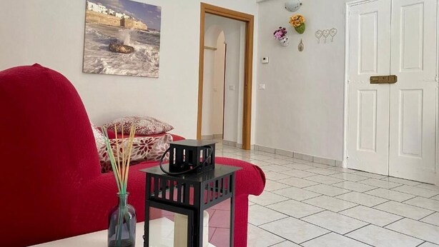 Un salon avec un canapé rouge, une petite table sur laquelle est posé un vase, un tableau représentant un paysage côtier et une porte blanche.