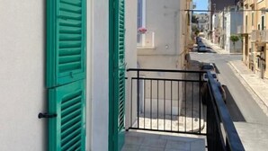 Une rue étroite, bordée de bâtiments des deux côtés, un balcon avec des volets verts et un sol carrelé.