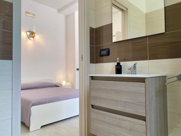 A modern bedroom with a bed, bedside table, and a bathroom with a vanity, mirror, and towel rack. - Sky (Monopoli)