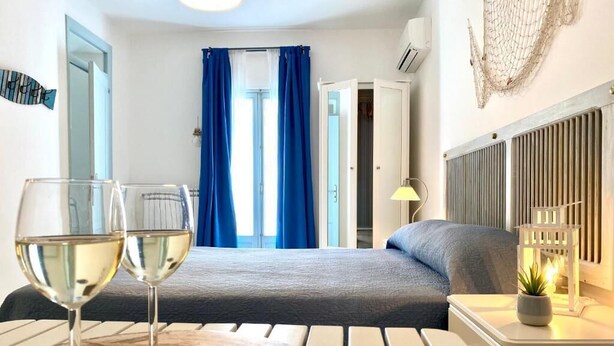 A room with a bed, a table with two glasses of white wine, a lamp, and a decorative wall piece. - Pearl of the Sea (Monopoli)