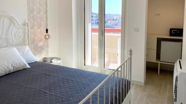 A bedroom with a bed, a nightstand, a window with a view, and a small desk with a computer. - Porto Bianco (Monopoli)
