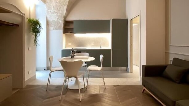 A modern interior with a herringbone wooden floor, a dining area with white chairs, a green sofa, and a kitchen with a dark green cabinet and a white countertop. - MARIACHIARA (Monopoli)