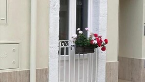 Un edificio de fachada blanca, con una ventana protegida por una barandilla metálica y una maceta con flores rojas y blancas.