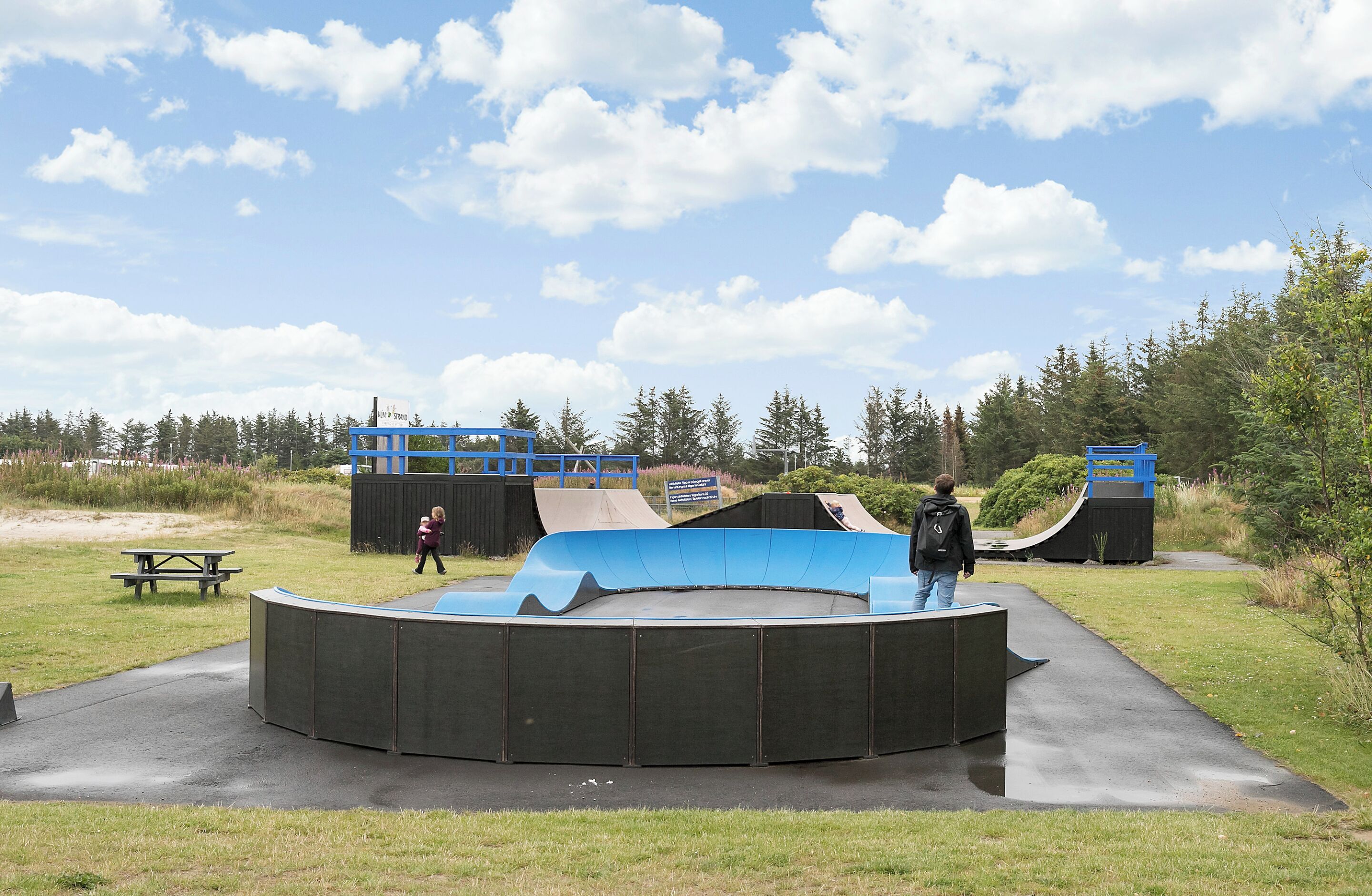 Ein Skatepark mit einer gebogenen Rampe, einem Picknicktisch und einer blauen Struktur im Hintergrund.