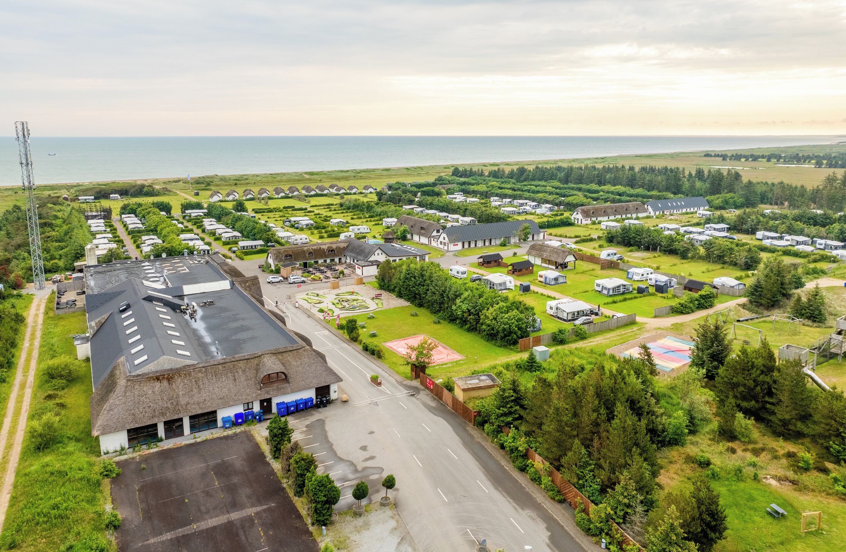 Vista aérea de un camping con vehículos recreativos y casas móviles, un edificio grande con techo de paja y vistas al mar.
