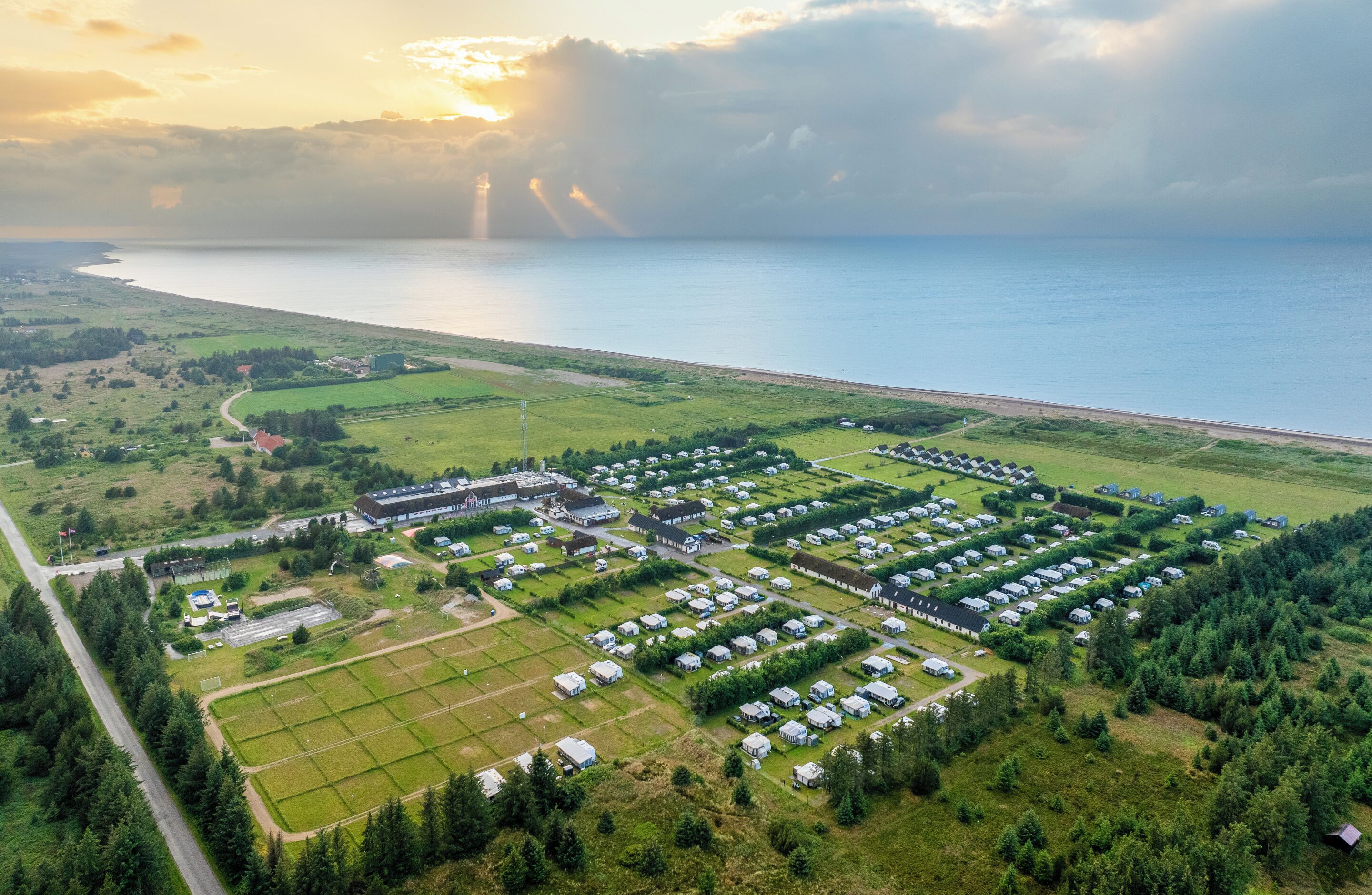 An aerial view of a coastal area with a large number of small, uniformly arranged cabins, a central building complex, and a road leading to the beach.