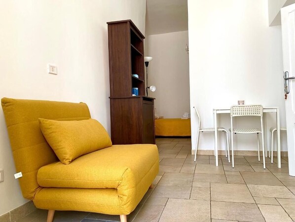 Une pièce avec un canapé jaune, un meuble en bois, une table blanche avec des chaises et un sol carrelé.