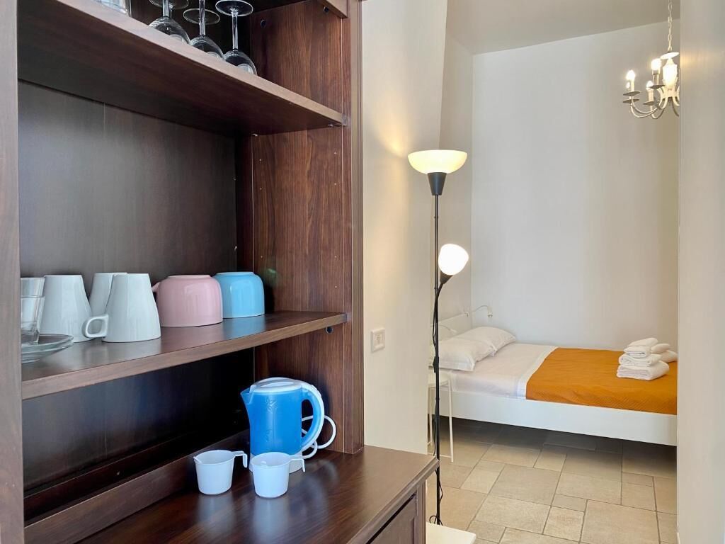 A compact room with a bed, a bedside table with cups and a kettle, a lamp, and a shelf with mugs and glasses.