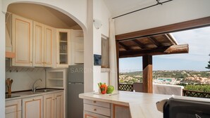 Eine Küche mit weißen Schränken, einer Spüle und einem Kühlschrank. Es gibt einen Tisch mit Blick auf eine Stadt und einen Balkon mit Holzboden.