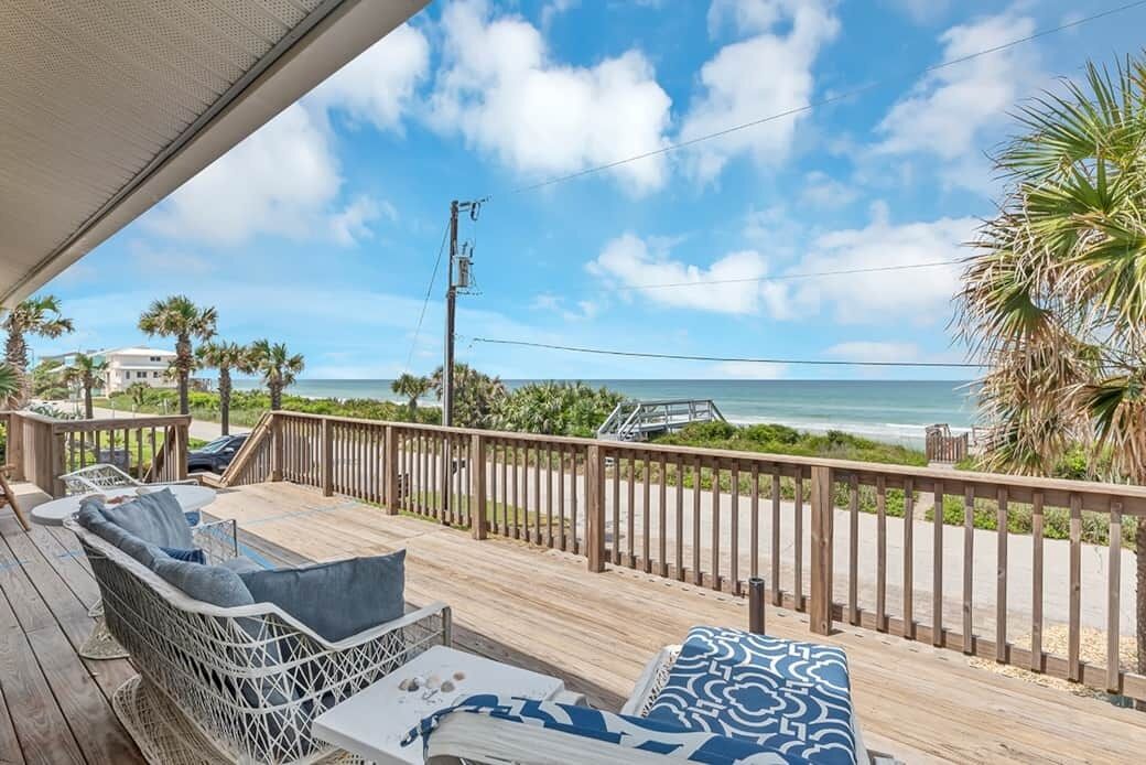 A wooden deck with a wicker sofa and a table, overlooking a beach with palm trees and a clear sky.