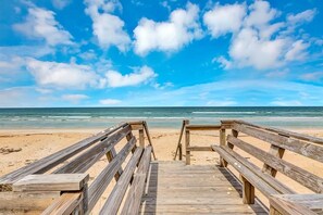 En træpromenade der fører ned til en strand med blå himmel og skyer.