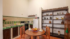 A compact kitchen with a dining area, featuring a round wooden table, chairs, a microwave, a refrigerator, and a shelf with kitchenware. - Casita Los Buhos (Antigua Guatemala)