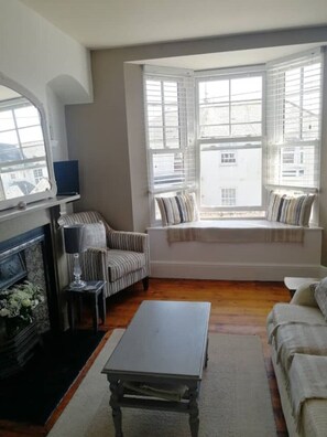 A living room with a fireplace, a striped armchair, a sofa, a window seat with pillows, and a view of buildings outside. - Gorgeous 3 bedroom home opposite beach. ! (Budleigh Salterton)