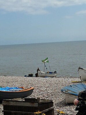 En strand med småsten och flera båtar, varav en har en flagga, och en person som står nära vattnet.