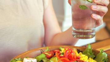 A person holding a glass of a clear beverage with lime and mint, and a bowl of colorful salad with shrimp, avocado, and corn.