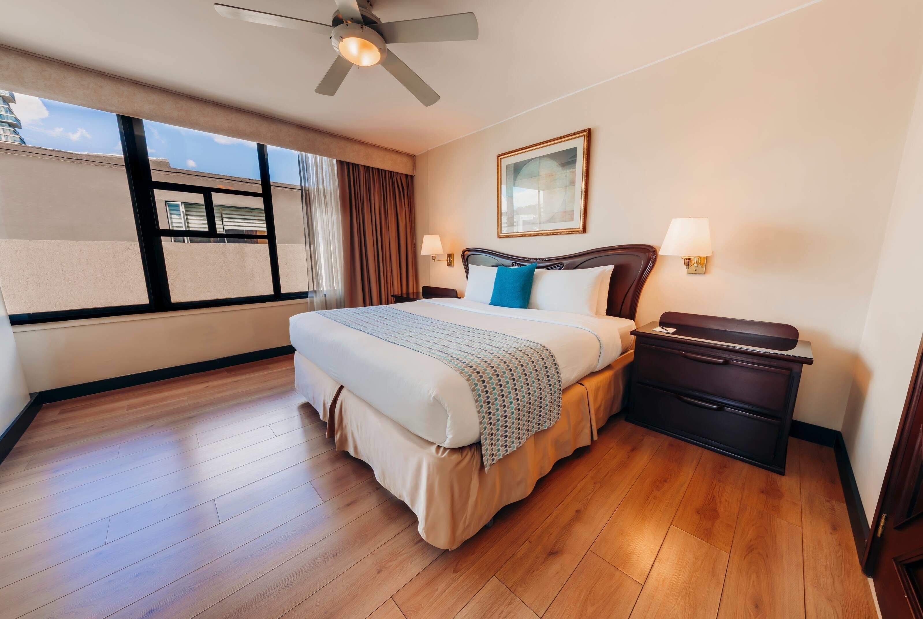 A hotel room with a bed, bedside tables, a ceiling fan, a window with curtains, and a wall-mounted lamp.