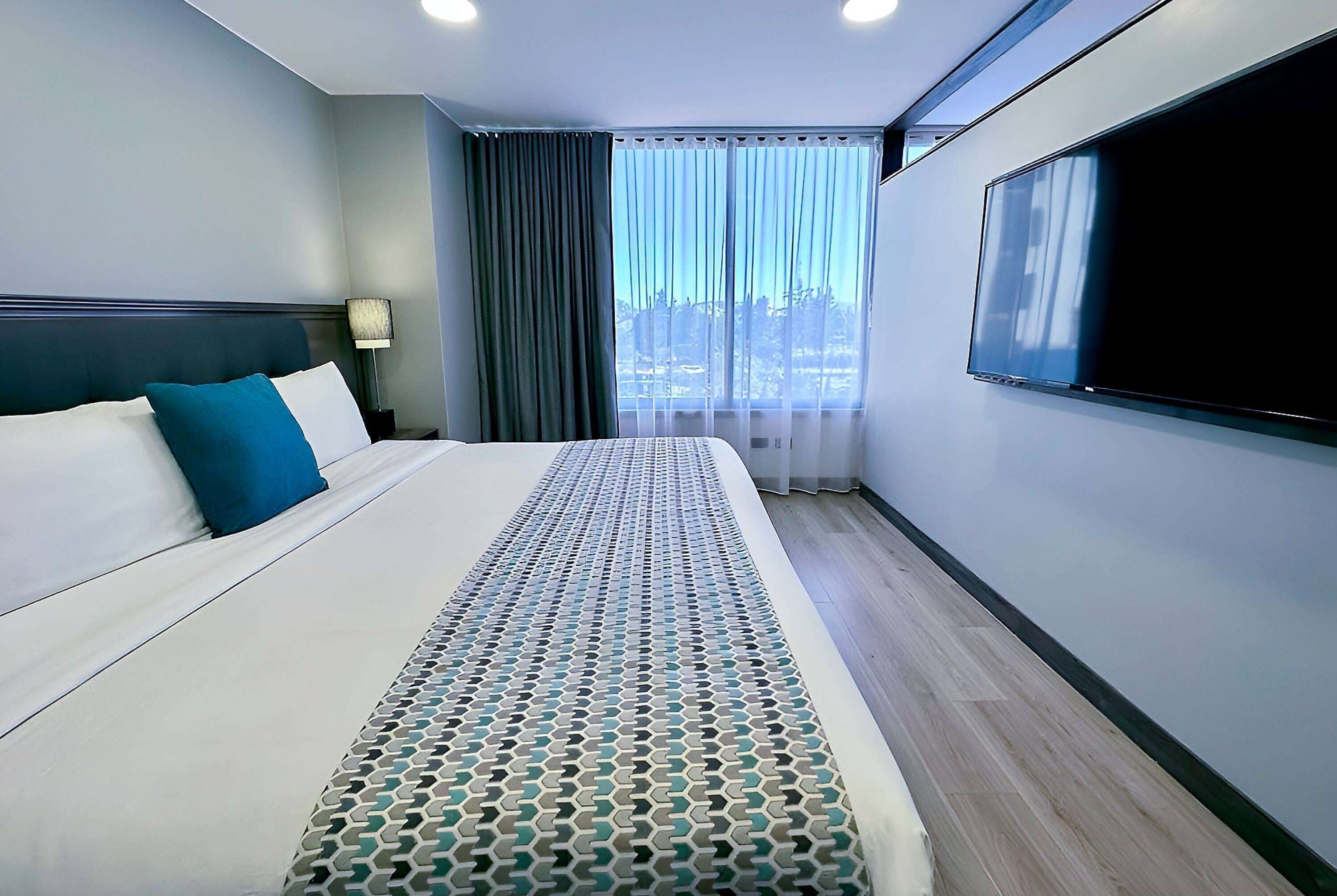 A hotel room with a large bed, a TV on the wall, and a window with curtains.