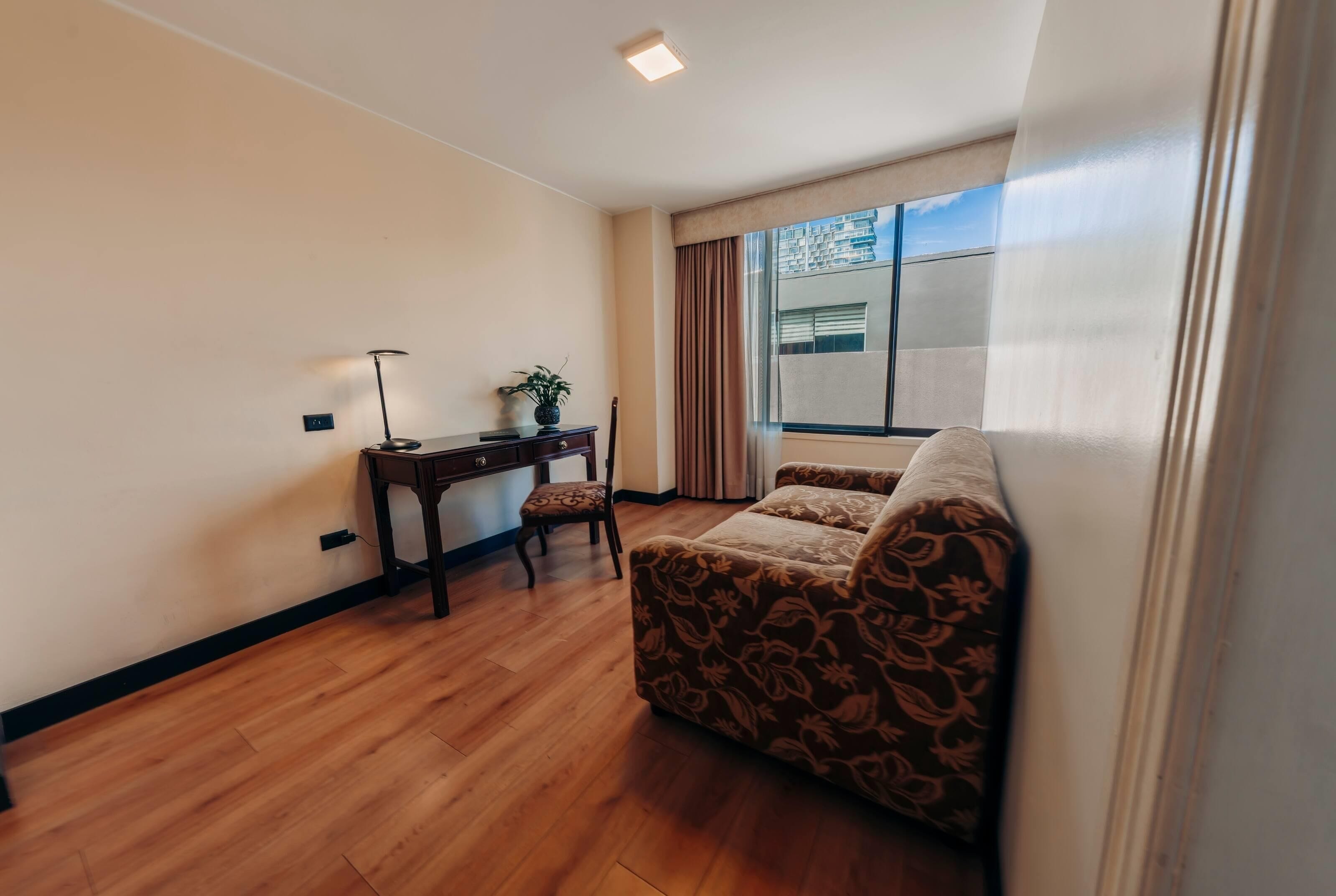 A room with a wooden floor, a sofa, a desk with a chair, and a window with curtains.