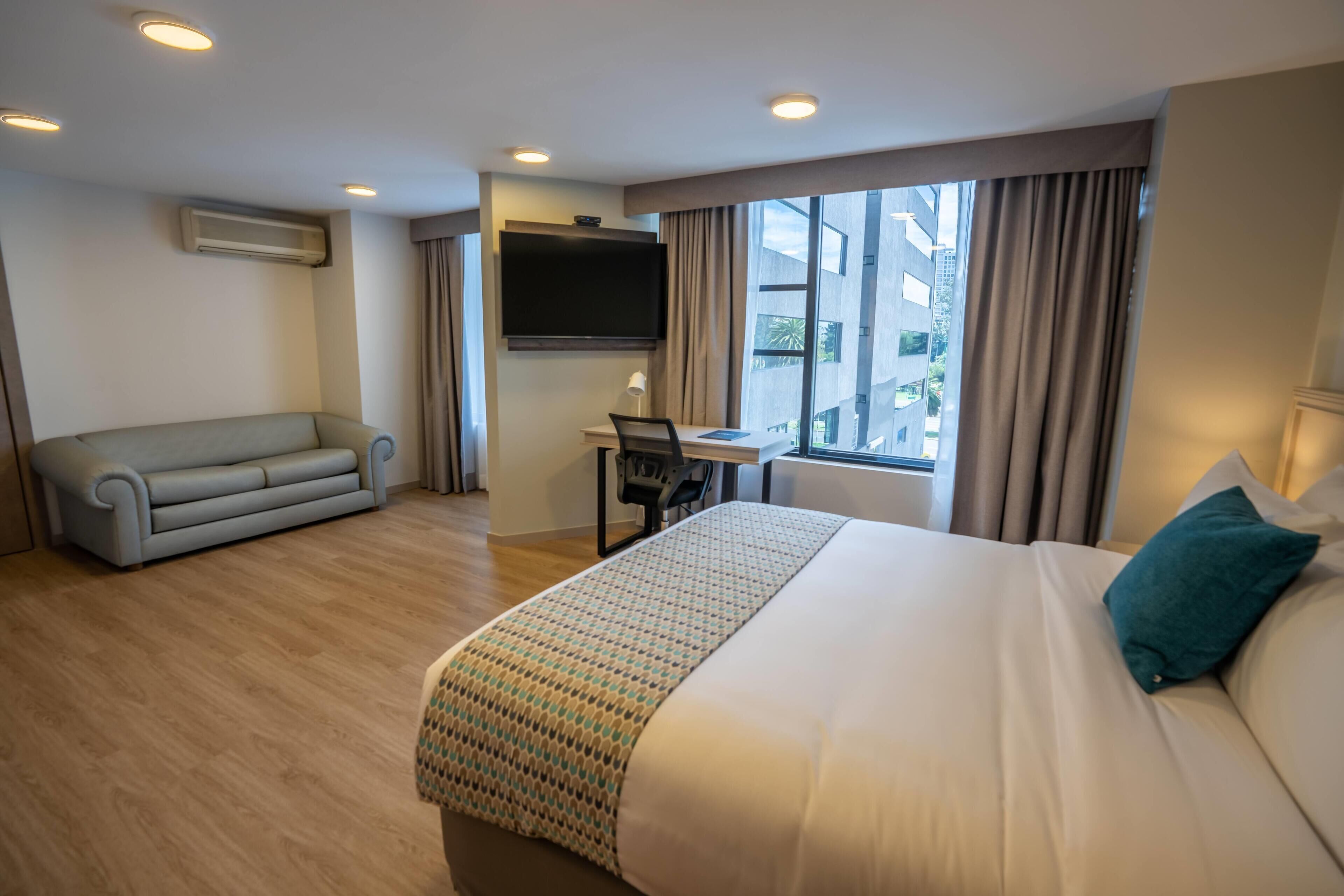 Habitación de hotel con una cama grande, un sofá, un escritorio con una silla, un televisor montado en la pared y una ventana con cortinas.