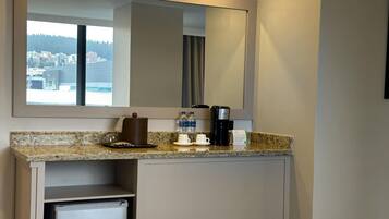 Baño de hotel con encimera de granito, cafetera, dos tazas y un refrigerador pequeño.