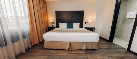 A hotel room with a large bed, wooden flooring, a dark headboard, two bedside tables with lamps, and a sliding glass door leading to a shower area.