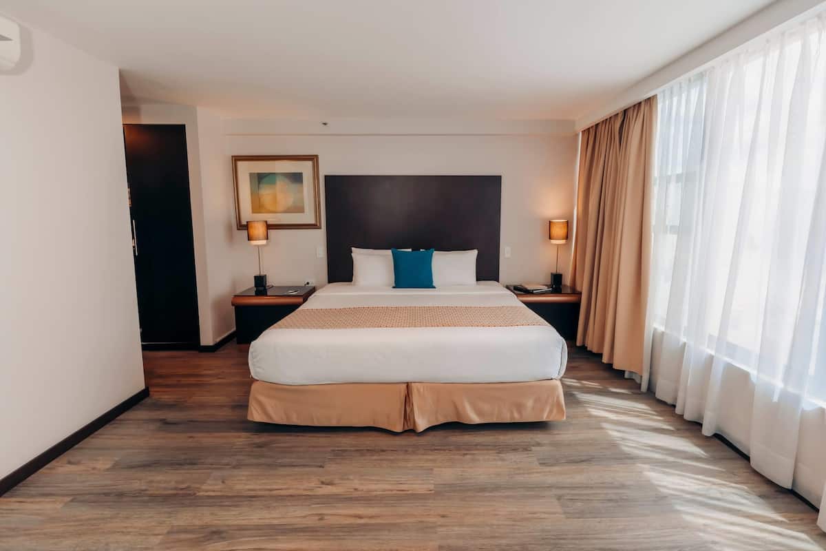A hotel room with a large bed, two bedside tables, a headboard, a painting, and a window with curtains.