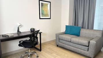Una sala moderna con un sofá gris, una silla de oficina negra, un escritorio con una lámpara y una obra de arte enmarcada en la pared.
