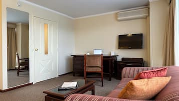Habitación de hotel con sofá, una mesa de centro, escritorio con computadora portátil, televisor de pantalla plana y puerta que da a otra habitación.