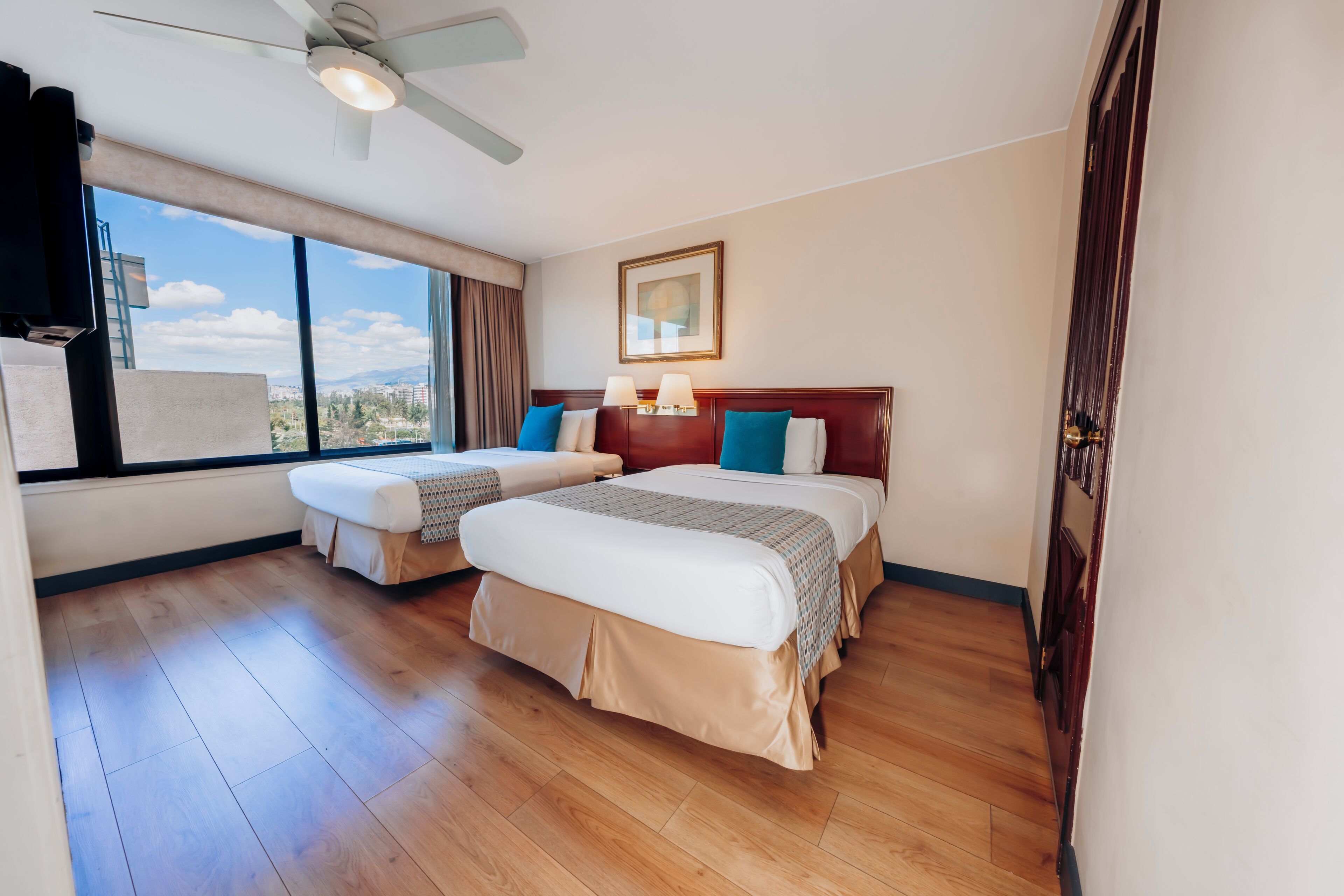 Une chambre d’hôtel avec deux lits, un ventilateur de plafond, une grande fenêtre donnant sur un beau paysage et un tableau encadré au mur.