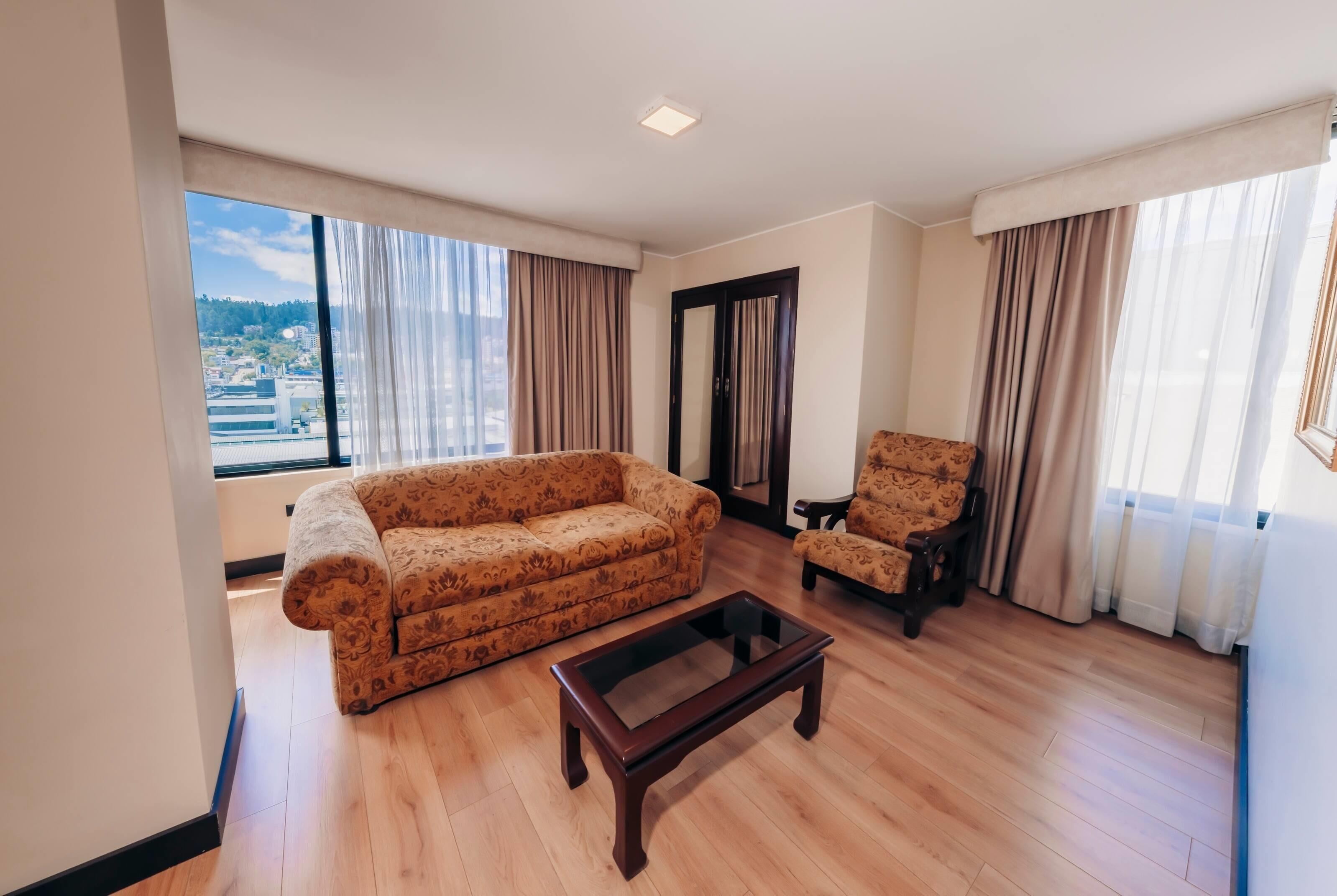 A living room with a sofa, armchair, and coffee table, large windows with curtains, and a view of a cityscape.